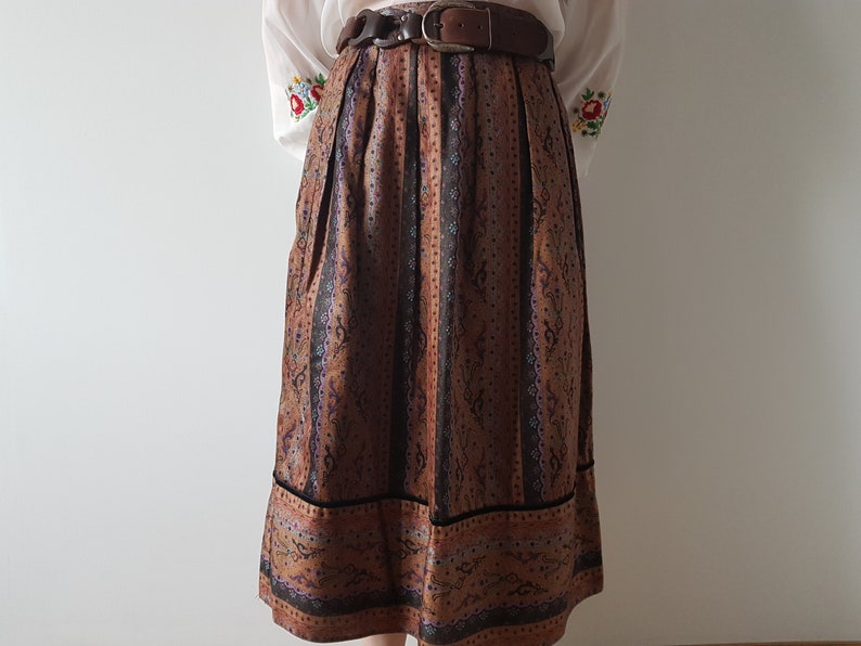 May include: A brown and black patterned skirt with a black velvet trim. The skirt has a high waist and is pleated. It is worn with a white blouse with embroidered flowers.