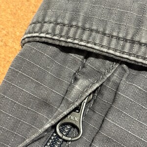 Pu&ograve; includere: Primo piano di tessuto grigio con trama e cuciture visibili. Una cerniera argentata con il marchio "YKK" &egrave; parzialmente aperta, rivelando i denti della cerniera. Il tessuto sembra far parte di un capo di abbigliamento, forse pantaloni o una giacca.