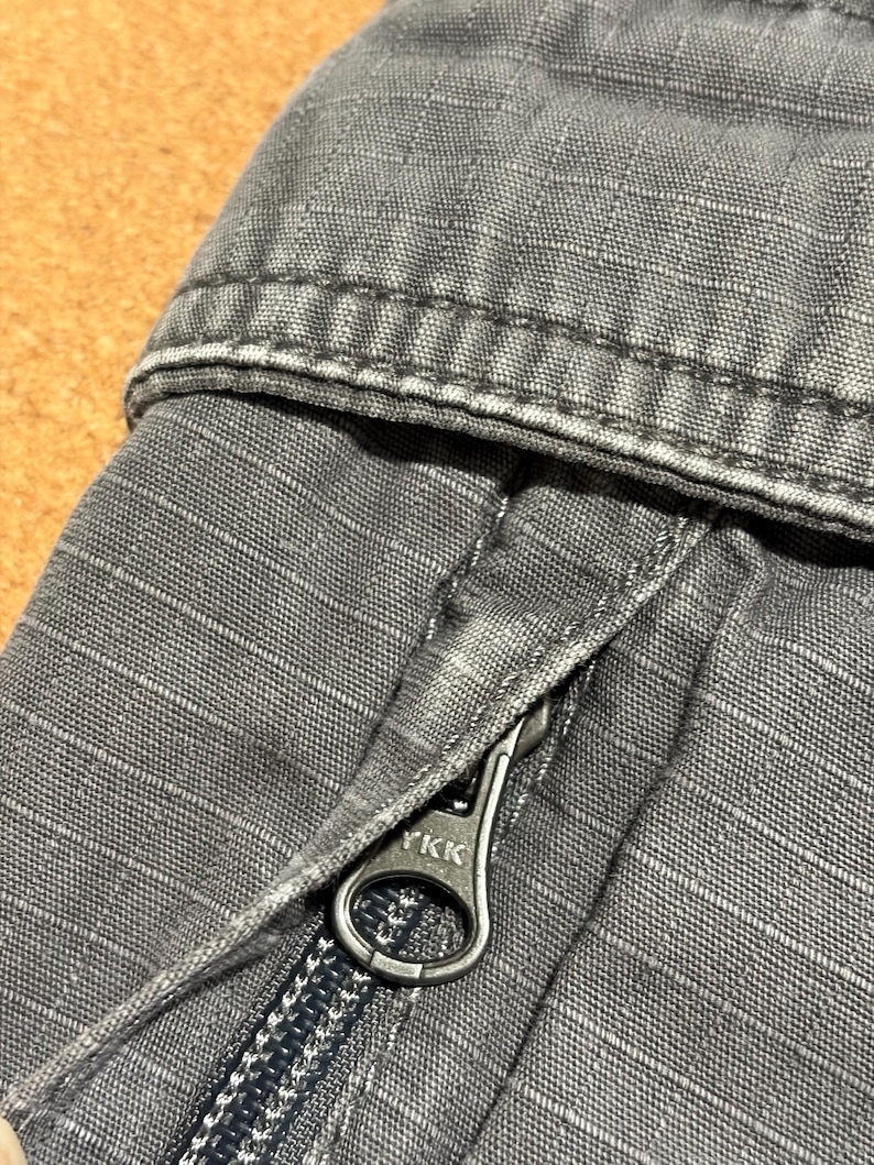 Pu&ograve; includere: Primo piano di tessuto grigio con trama e cuciture visibili. Una cerniera argentata con il marchio "YKK" &egrave; parzialmente aperta, rivelando i denti della cerniera. Il tessuto sembra far parte di un capo di abbigliamento, forse pantaloni o una giacca.