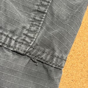 Pu&ograve; includere: Primo piano di tessuto grigio con cuciture visibili. Il tessuto presenta un motivo strutturato a griglia. Le cuciture sono grigio scuro e sembrano rinforzate. L'immagine evidenzia i dettagli del tessuto e della costruzione.