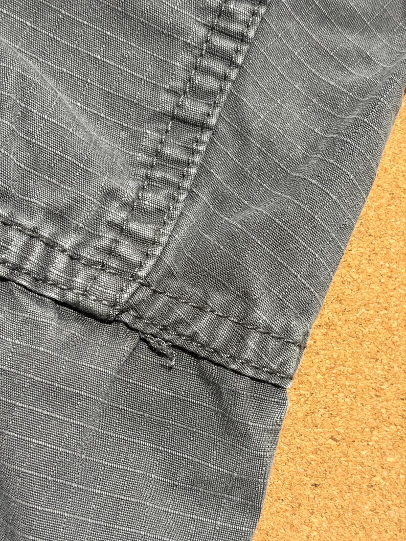 Pu&ograve; includere: Primo piano di tessuto grigio con cuciture visibili. Il tessuto presenta un motivo strutturato a griglia. Le cuciture sono grigio scuro e sembrano rinforzate. L'immagine evidenzia i dettagli del tessuto e della costruzione.