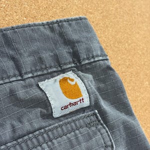 Pu&ograve; includere: Primo piano di pantaloni da lavoro Carhartt grigi. La toppa con il logo Carhartt &egrave; visibile, con un emblema dorato e il nome del marchio in rosso. I pantaloni hanno un tessuto strutturato e un dettaglio tasca.