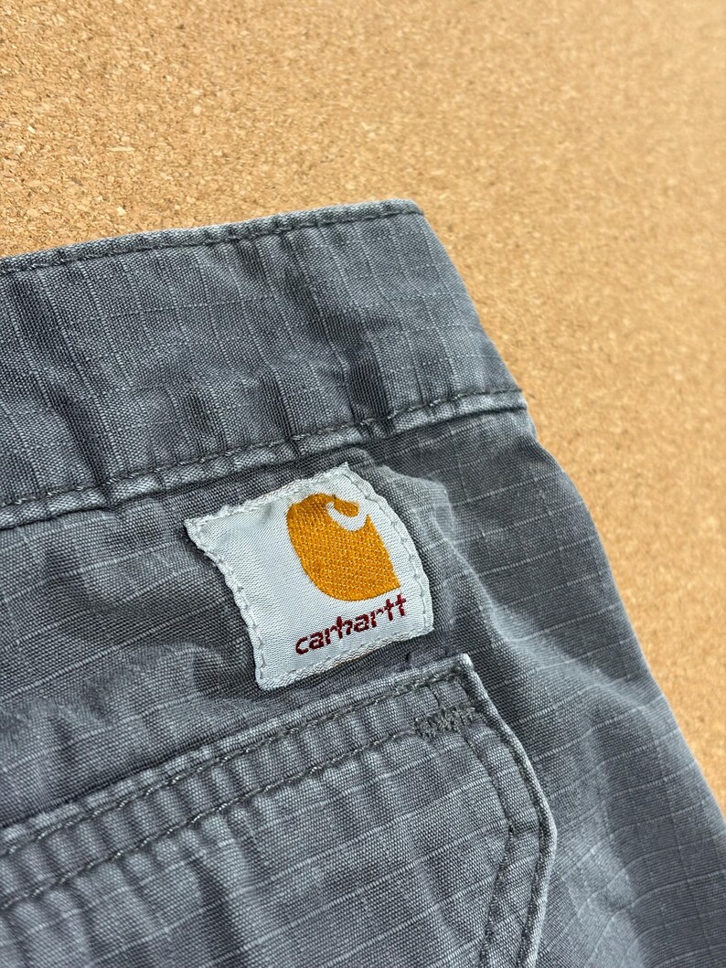 Pu&ograve; includere: Primo piano di pantaloni da lavoro Carhartt grigi. La toppa con il logo Carhartt &egrave; visibile, con un emblema dorato e il nome del marchio in rosso. I pantaloni hanno un tessuto strutturato e un dettaglio tasca.