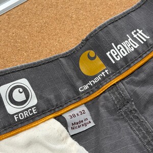Pu&ograve; includere: Primo piano di pantaloni da lavoro grigi Carhartt con vestibilit&agrave; rilassata. Il logo Carhartt &egrave; visibile, insieme alla parola "FORCE" e alla taglia 38x32. I pantaloni sono fabbricati in Nicaragua.