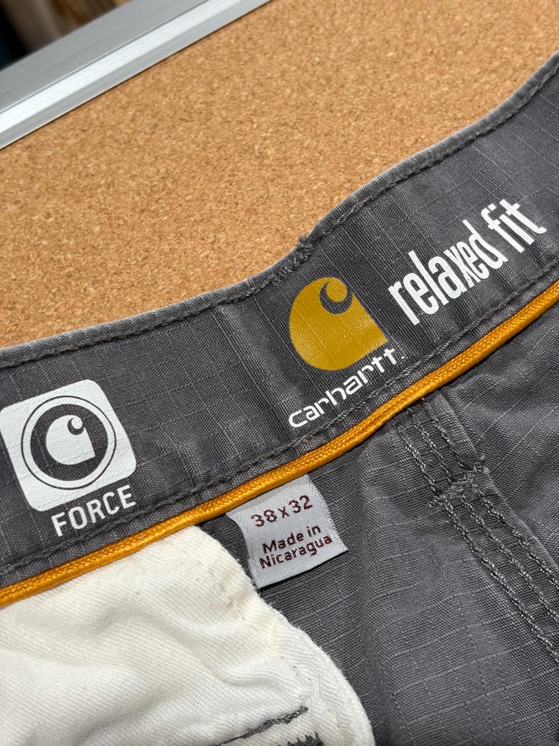 Pu&ograve; includere: Primo piano di pantaloni da lavoro grigi Carhartt con vestibilit&agrave; rilassata. Il logo Carhartt &egrave; visibile, insieme alla parola "FORCE" e alla taglia 38x32. I pantaloni sono fabbricati in Nicaragua.