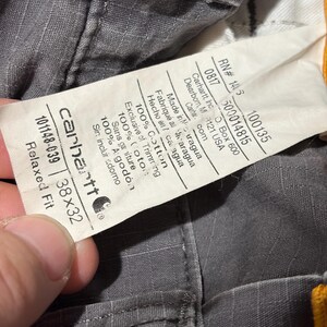 Pu&ograve; includere: Primo piano di pantaloni Carhartt grigi con un'etichetta che mostra il logo del marchio, la taglia 96,5x81,3 cm e i dettagli del tessuto. L'etichetta include anche il numero RN e lo stile. I pantaloni sono in 100% cotone.