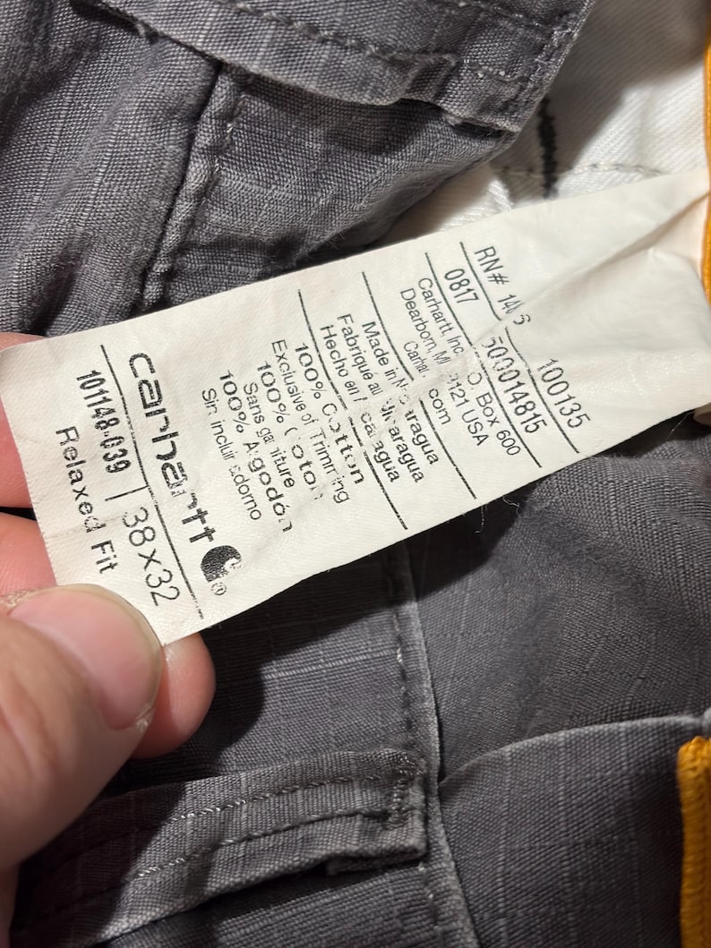 Pu&ograve; includere: Primo piano di pantaloni Carhartt grigi con un'etichetta che mostra il logo del marchio, la taglia 96,5x81,3 cm e i dettagli del tessuto. L'etichetta include anche il numero RN e lo stile. I pantaloni sono in 100% cotone.