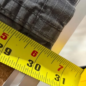 Pu&ograve; includere: Primo piano di tessuto grigio con cuciture visibili. Un metro a nastro giallo mostra una misura di 76 cm. Il tessuto sembra far parte di un capo di abbigliamento, possibilmente pantaloni o pantaloncini.