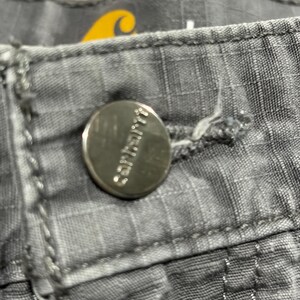 Pu&ograve; includere: Primo piano di pantaloni Carhartt grigi con un bottone argentato. Il bottone &egrave; impresso con il logo Carhartt. I pantaloni hanno un motivo strutturato a griglia e cuciture visibili. Un logo giallo &egrave; visibile sullo sfondo.