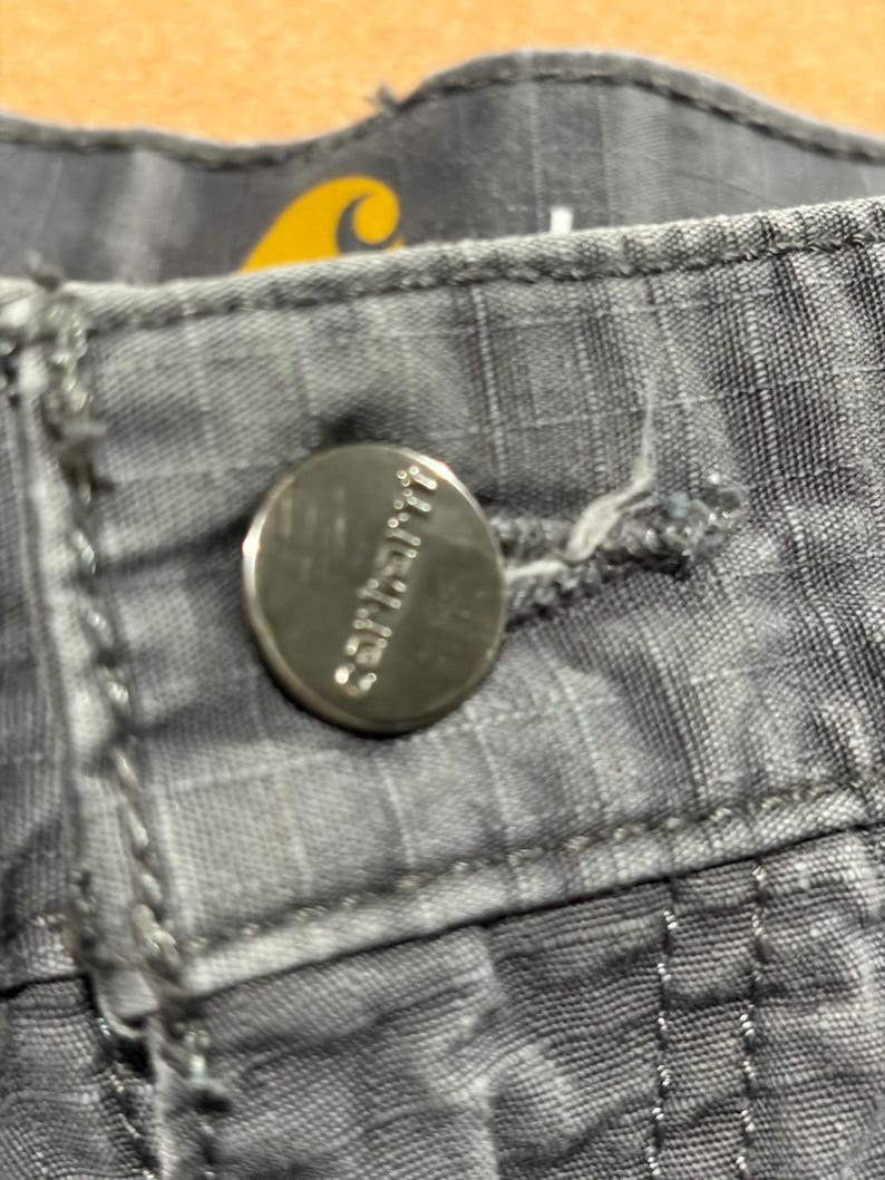 Pu&ograve; includere: Primo piano di pantaloni Carhartt grigi con un bottone argentato. Il bottone &egrave; impresso con il logo Carhartt. I pantaloni hanno un motivo strutturato a griglia e cuciture visibili. Un logo giallo &egrave; visibile sullo sfondo.