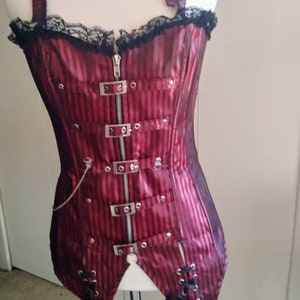 May include: A burgundy and black corset with vertical stripes, a front zipper, and silver-colored buckles. The corset has black lace trim and is displayed on a mannequin. A black lace choker with a red rose and pendant is also visible.
