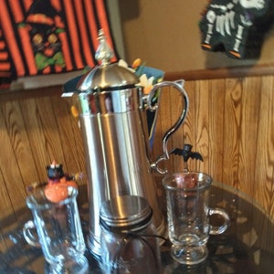 May include: A silver coffee press, a glass jar with a silver lid, and two clear glass mugs are on a glass table. A small black bat decoration is on one of the mugs. The scene has a Halloween theme.