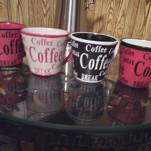 May include: Four ceramic coffee mugs with the word "Coffee" in large letters. The mugs are red, white, and black with the words "Coffee" and "BREAK" printed on them. The mugs are sitting on a glass table.