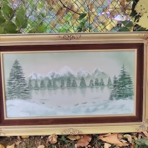 May include: A framed landscape painting depicting a snowy mountain scene with evergreen trees and a reflective lake. The painting is encased in a gold-colored ornate frame with a dark brown inner border.