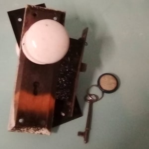 May include: An antique door lock set with a white ceramic doorknob, a key, and a key ring. The lock plate is dark brown with orange and white accents. The key is silver-colored.