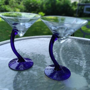 May include: Two clear martini glasses with a unique design. The stems are a deep blue, curving gracefully from the base to support the glass bowl. The glasses are on a glass table, with a blurred green background.