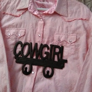 May include: Pink button-down shirt with two chest pockets and a collar. A black wooden sign that says "COWGIRL" is attached to the shirt. The shirt has a Western style.