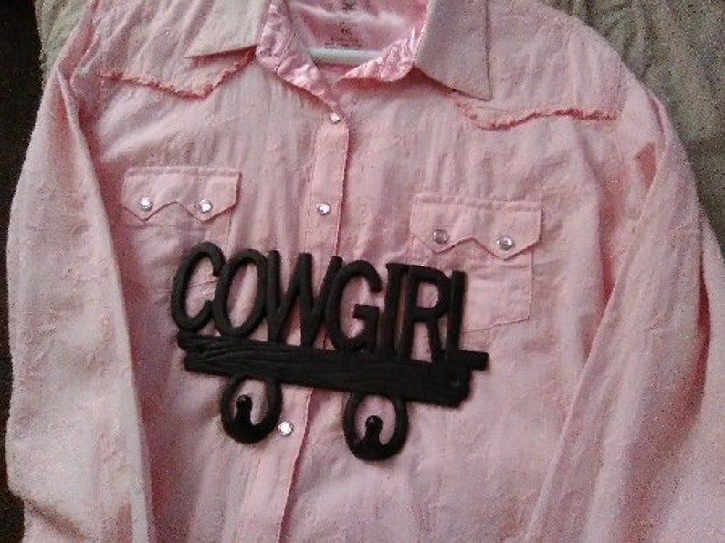 May include: Pink button-down shirt with two chest pockets and a collar. A black wooden sign that says "COWGIRL" is attached to the shirt. The shirt has a Western style.