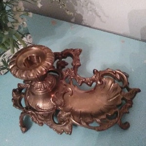 May include: Ornate antique brass candle holder with a decorative, scrollwork design. The candle holder features a circular base with a detailed floral pattern and a small dish. A vintage home decor item.