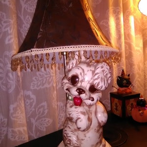 May include: A vintage ceramic dog lamp with a brown lampshade. The dog has a red tongue and a green collar. The lampshade is brown with a decorative trim and hanging beads. A Halloween-themed cat figurine is in the background.