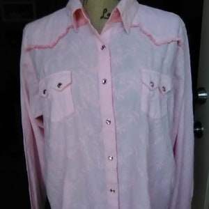 May include: A light pink button-down shirt with long sleeves. The shirt has two chest pockets and silver-colored buttons. The collar and yoke feature a decorative pink trim. The fabric has a subtle pattern.