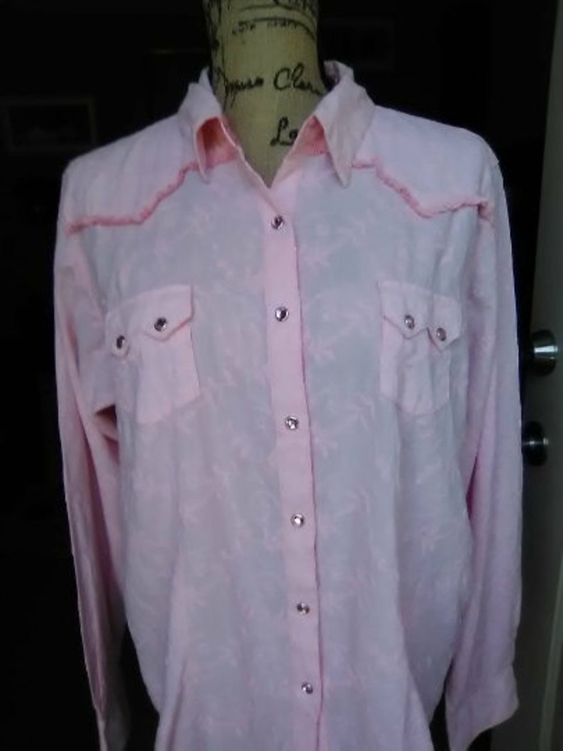 May include: A light pink button-down shirt with long sleeves. The shirt has two chest pockets and silver-colored buttons. The collar and yoke feature a decorative pink trim. The fabric has a subtle pattern.