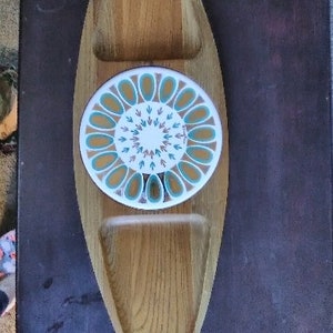 May include: Wooden serving tray with a central circular ceramic insert. The insert features a white background with a teal and orange floral design. The tray has two recessed sections and is made of light-colored wood.