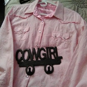 May include: A light pink button-down shirt with two chest pockets and a collar. The shirt has a decorative black "COWGIRL" sign attached to the front. The shirt is hanging on a white hanger.