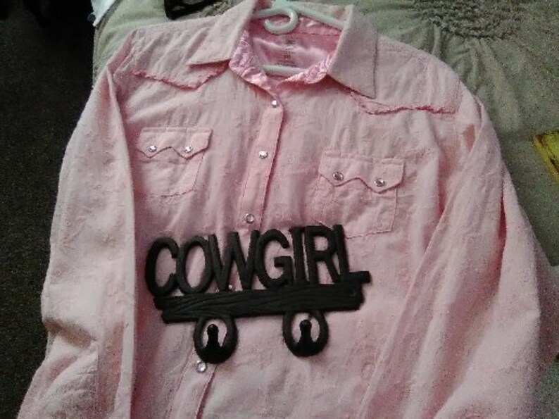May include: A light pink button-down shirt with two chest pockets and a collar. The shirt has a decorative black "COWGIRL" sign attached to the front. The shirt is hanging on a white hanger.