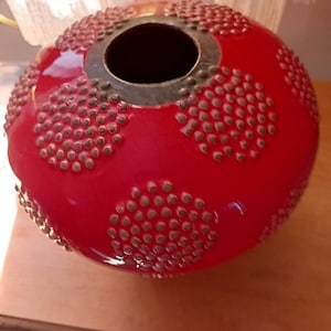 May include: A round, red decorative vase with a glossy finish. The vase features a series of raised, bronze-colored dots arranged in circular patterns across its surface. The top has a dark, circular opening with a bronze-colored rim.