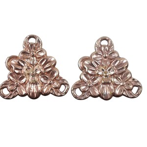 May include: Two gold-tone metal flower-shaped charms with a triangular shape. Each charm has three flowers with a raised design and a small hole at the top.