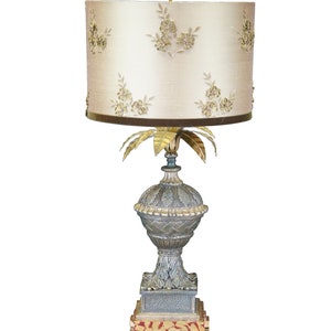 May include: A gold and blue table lamp with a floral patterned lampshade. The lamp base is a stylized urn with a palm tree design.