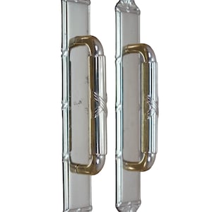May include: Two vertical door handles featuring a rectangular silver-toned backplate with decorative embossed ends. Each handle has a central gold-toned grip section with a cross-pattern detail, flanked by silver-toned ends, mounted on the plate.