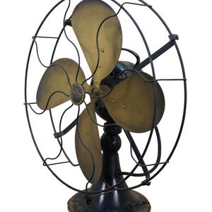 Puede incluir: Un ventilador el&eacute;ctrico Emerson antiguo con cuatro aspas de lat&oacute;n y un marco y base de metal negro. El ventilador tiene una jaula circular y una base decorativa, mostrando un dise&ntilde;o vintage.