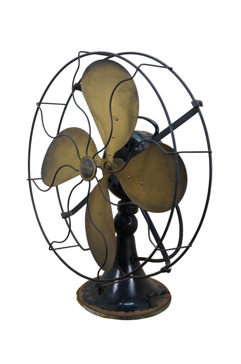 Puede incluir: Un ventilador el&eacute;ctrico Emerson antiguo con cuatro aspas de lat&oacute;n y un marco y base de metal negro. El ventilador tiene una jaula circular y una base decorativa, mostrando un dise&ntilde;o vintage.