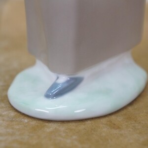 May include: Close-up of a ceramic figurine base. The base is a light gray color with a white and light green oval shape. A small, blue-gray accent is visible on the base. The figurine appears to be a decorative item.