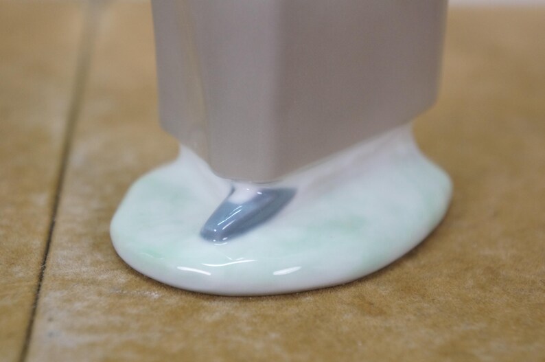 May include: Close-up of a ceramic figurine base. The base is a light gray color with a white and light green oval shape. A small, blue-gray accent is visible on the base. The figurine appears to be a decorative item.