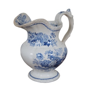 May include: A white ceramic pitcher with a blue floral pattern. The pitcher has a wide mouth and a handle.