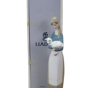May include: A porcelain figurine of a woman holding a lamb, with a box in the background. The woman wears a blue top and a long white and brown skirt. The lamb is white. The box has the word "LLAD" printed on it.