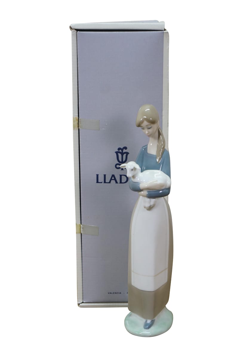 May include: A porcelain figurine of a woman holding a lamb, with a box in the background. The woman wears a blue top and a long white and brown skirt. The lamb is white. The box has the word "LLAD" printed on it.