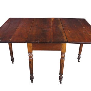 May include: A rectangular, antique wooden drop-leaf table with a dark brown finish. The table has four turned legs with small wheels and a central drawer. The leaves are extended, creating a larger surface area.