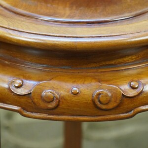 May include: Close-up of a wooden, antique plant stand. The stand is a warm, medium brown color with a glossy finish. The stand features decorative, carved cloud-like designs and a circular top.