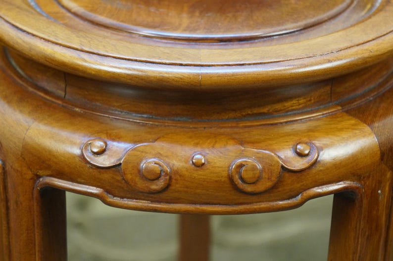 May include: Close-up of a wooden, antique plant stand. The stand is a warm, medium brown color with a glossy finish. The stand features decorative, carved cloud-like designs and a circular top.