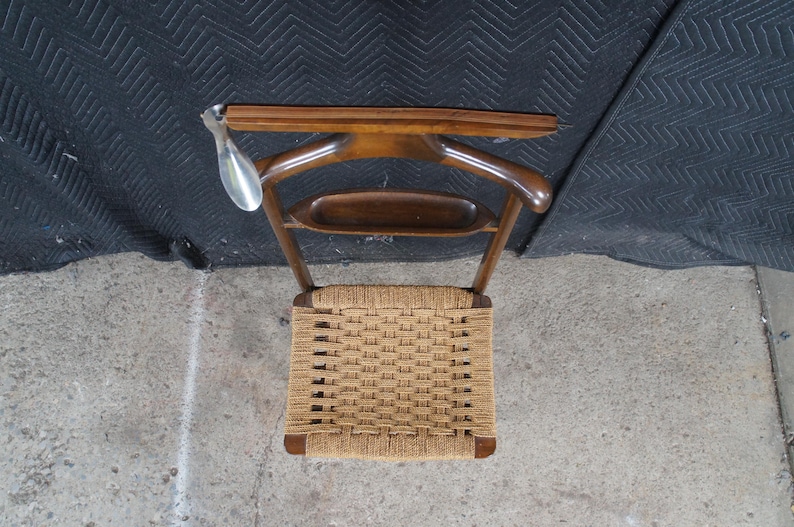 May include: A vintage wooden valet chair with a woven seat. The chair has a curved back and a bar for hanging clothes. A metal shoe horn hangs from the top. The chair is made of dark wood.