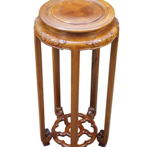 May include: A wooden plant stand with a round top and a decorative, openwork base. The stand is made of a warm-toned wood and features carved details around the top and at the base of the legs. The stand is approximately 20 inches tall.