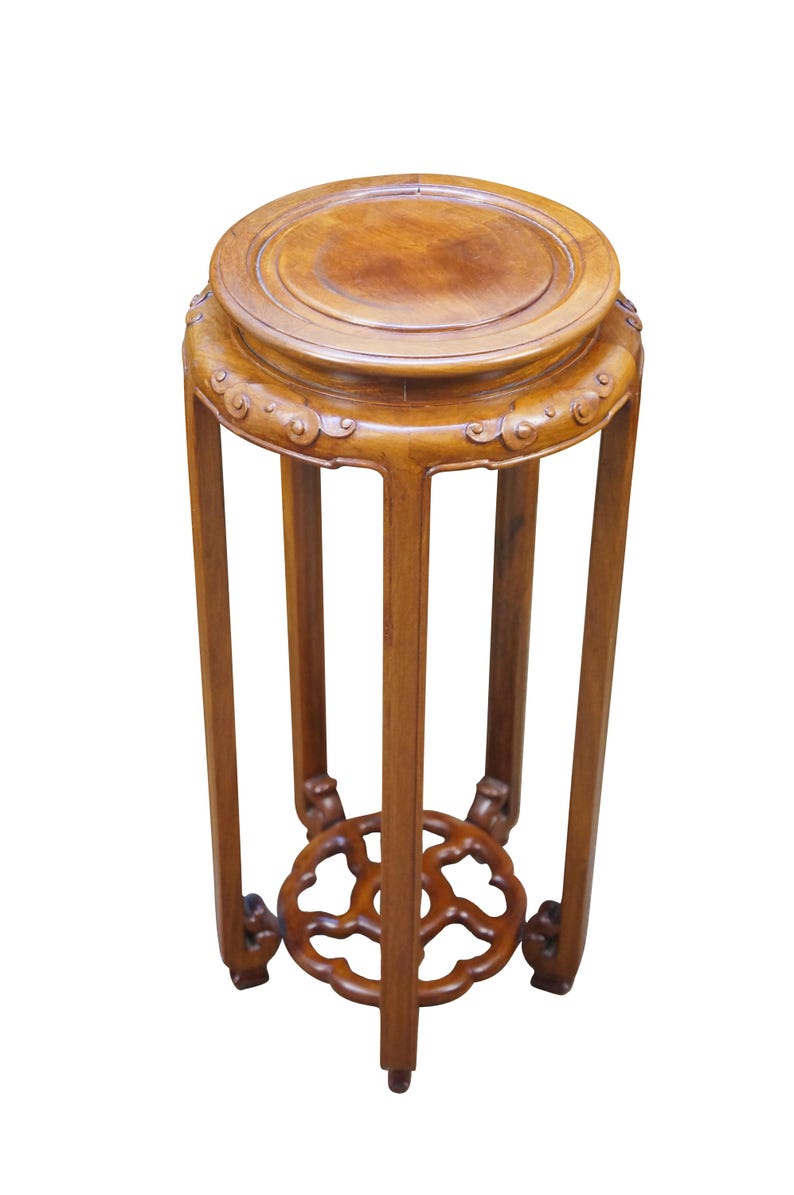 May include: A wooden plant stand with a round top and a decorative, openwork base. The stand is made of a warm-toned wood and features carved details around the top and at the base of the legs. The stand is approximately 20 inches tall.