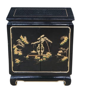 May include: Black lacquered cabinet with gold trim and decorative landscape art. The cabinet features two doors and a top with a subtle geometric pattern. The landscape art includes trees, buildings, and foliage.