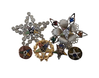 May include: A collection of five Masonic pins featuring a variety of designs, including a star with rhinestones, a star with a rose, a wreath with a star and the number 50, a star with a lantern, and a star with a compass and square.