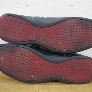May include: Pair of black athletic shoes with red soles. The soles have a textured pattern of small, circular indentations. The shoes appear worn, with some scuff marks visible. The uppers are black, and the shoes are shown from the bottom.