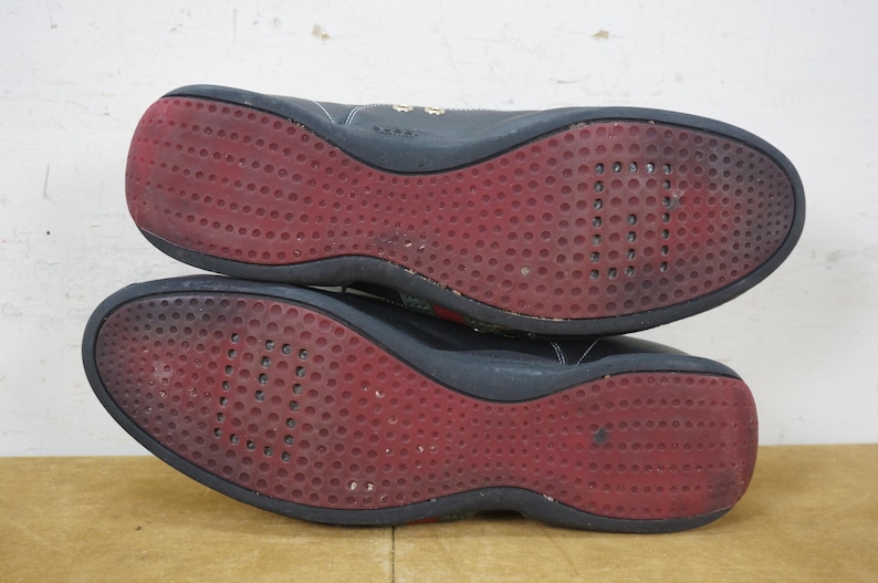 May include: Pair of black athletic shoes with red soles. The soles have a textured pattern of small, circular indentations. The shoes appear worn, with some scuff marks visible. The uppers are black, and the shoes are shown from the bottom.